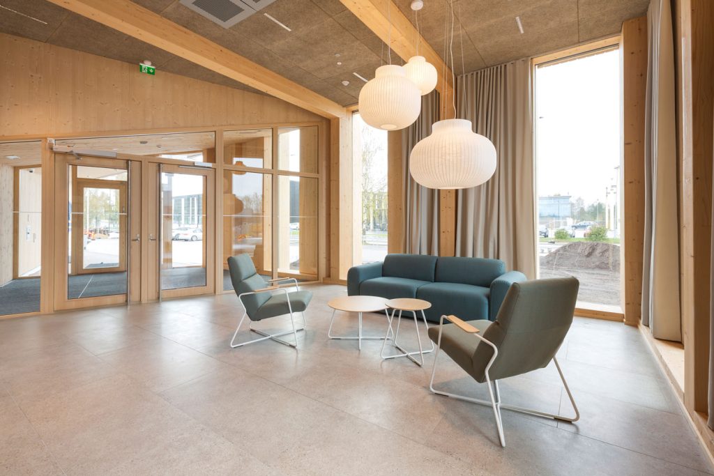 Rakennuksen aula, jossa on sohva, nojatuoleja ja pienet pöydät suurten ikkunoiden ja riippuvalaisimien äärellä.