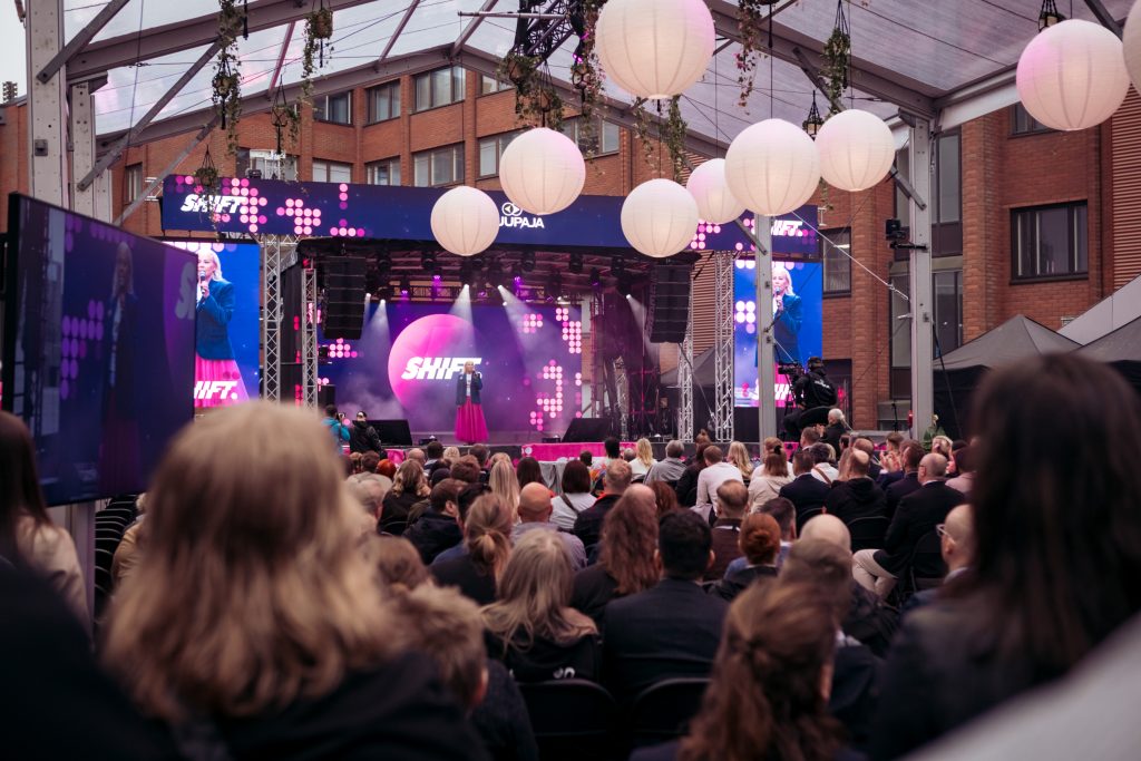 Yleisö seuraa puhujaa SHIFT businessfestivaalin päälavalla katetussa ulkotilassa, lavalla puhuja ja taustalla suuret näyttöpinnat ja valaistus.