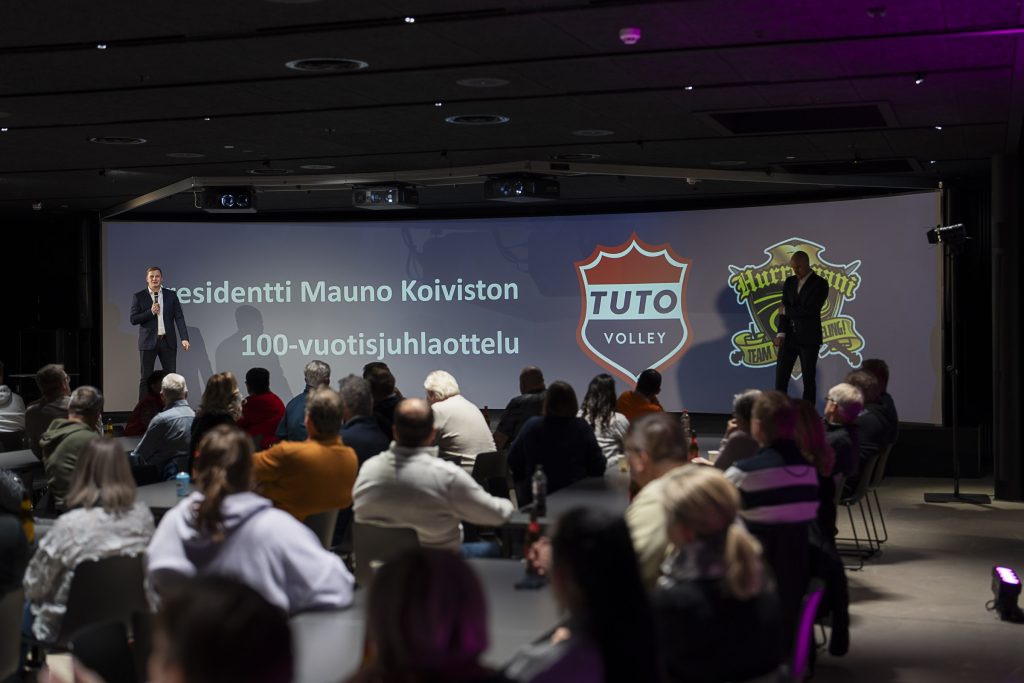 Yleisö seuraa esitystä tapahtumatilassa, lavalla puhuja ja taustalla suuri näyttö, jossa tapahtuman otsikko ja logot.