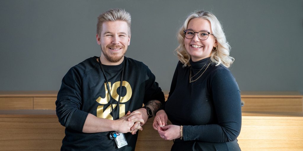 Kaksi henkilöä nojaa korkeaan pöytä- tai tiski­rakenteeseen sisätilassa, taustalla yksivärinen seinä.