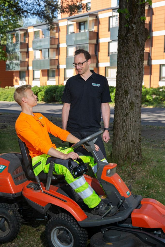 Kaksi henkilöä keskustelee ulkona viheralueella istuttavan ajoleikkurin vieressä, taustalla puu ja asuinrakennuksia.