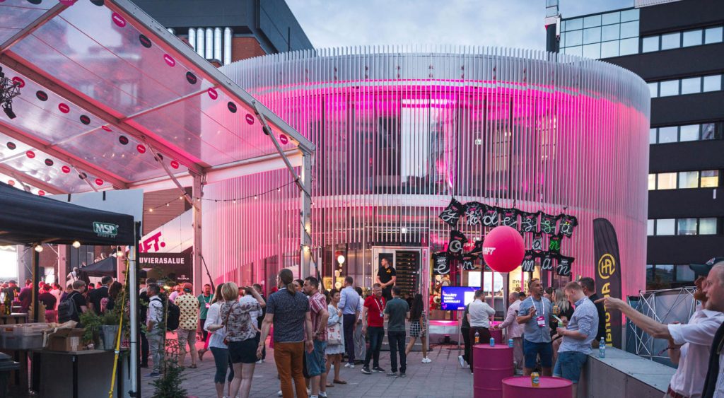 Människor pratar och rör sig på evenemangsområdet under SHIFT-businessfestivalen, med en upplyst rund byggnad och evenemangskonstruktioner i bakgrunden.
