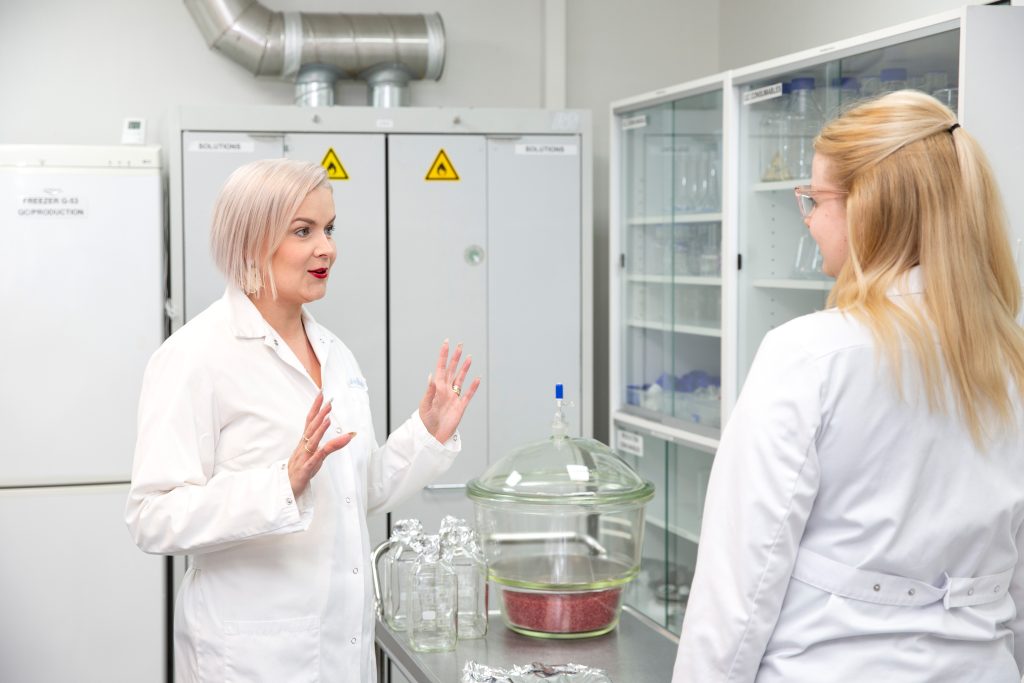 Kaksi henkilöä keskustelee laboratoriotilassa työpöydän äärellä, ympärillä laboratoriolaitteita ja suojakaapit.