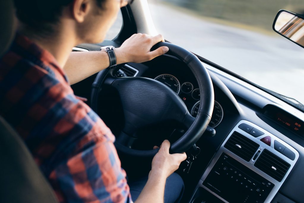 Henkilö ajaa autoa, näkymä ohjauspyörän ja kojelaudan yli tielle sisältäpäin kuvattuna.