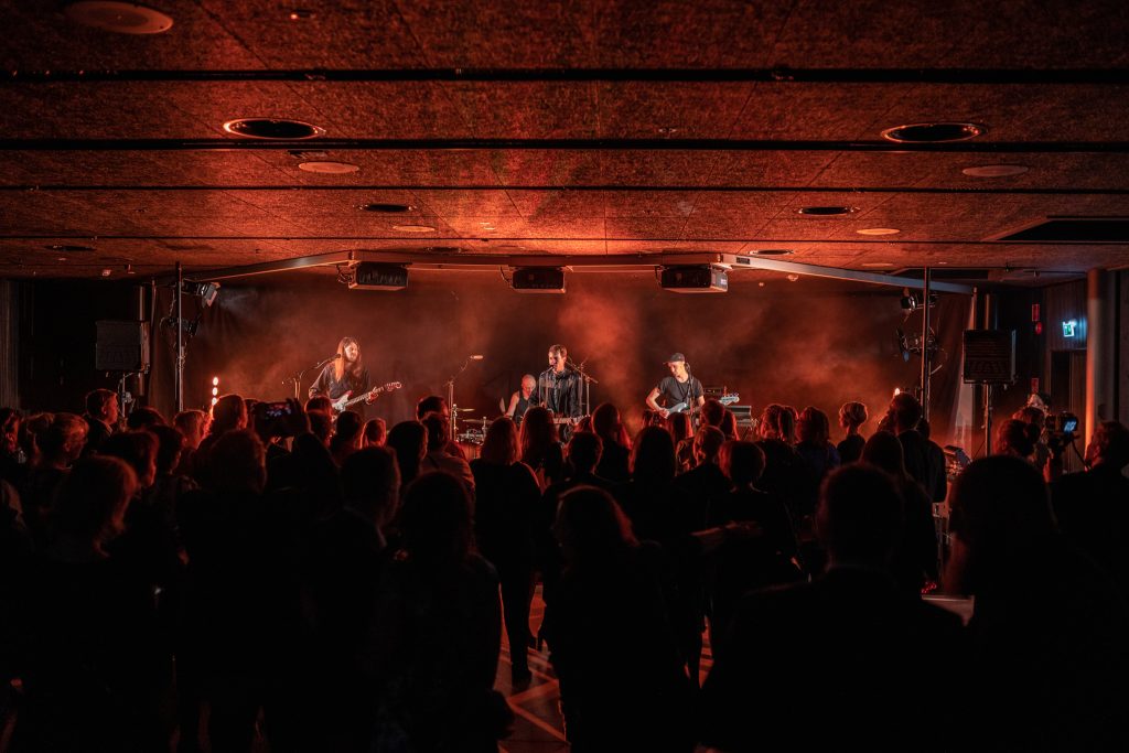 Elävä musiikkiesitys sisätilassa, lavalla esiintyjiä ja edessä seisova yleisö valaistussa tapahtumatilassa.