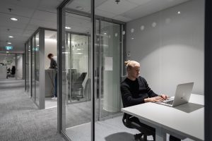 En person arbetar vid en bärbar dator i ett kontorsrum med glasväggar, med en lång korridor och återkommande arbetsplatser i bakgrunden.