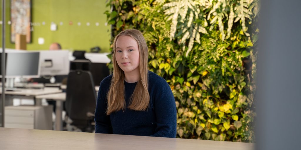 Henkilö istuu pöydän ääressä toimistotilassa, taustalla viherseinä ja avoimia työpisteitä.
