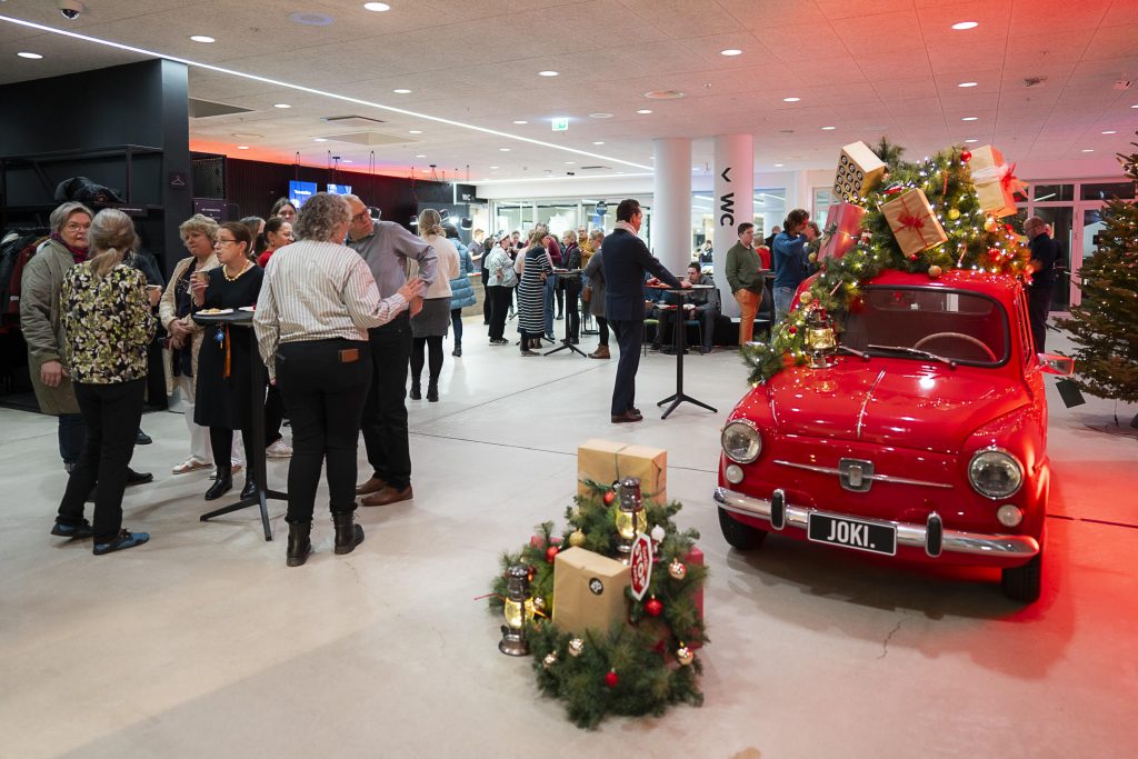 Aulatila tapahtuman aikana, ihmiset keskustelevat korkeiden pöytien äärellä ja etualalla joulukoristein somistettu punainen auto.