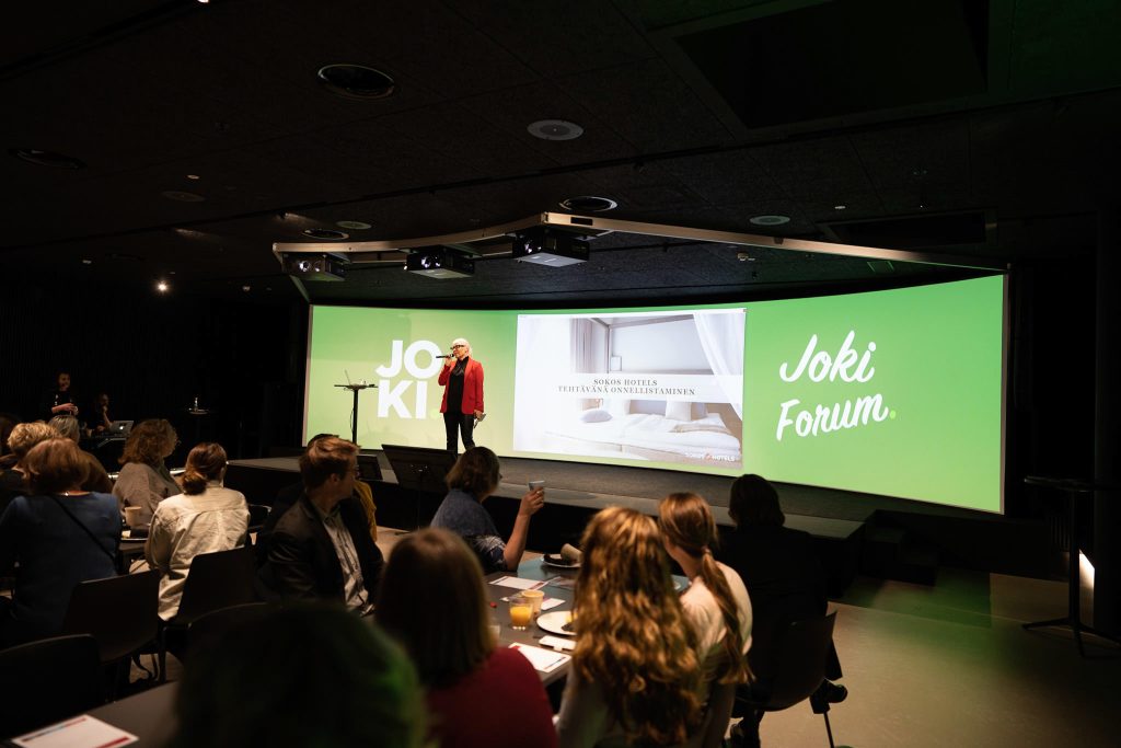 Esitys käynnissä Jeon Cavessa, yleisö istuu pöytien äärellä ja lavalla on puhuja suuren näytön edessä.