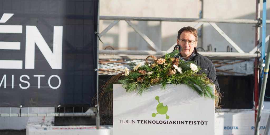 Henkilö puhuu mikrofonin ääressä ulkotilaisuudessa, edessä kukkakoristeinen puhujapönttö ja taustalla rakennustelineitä.