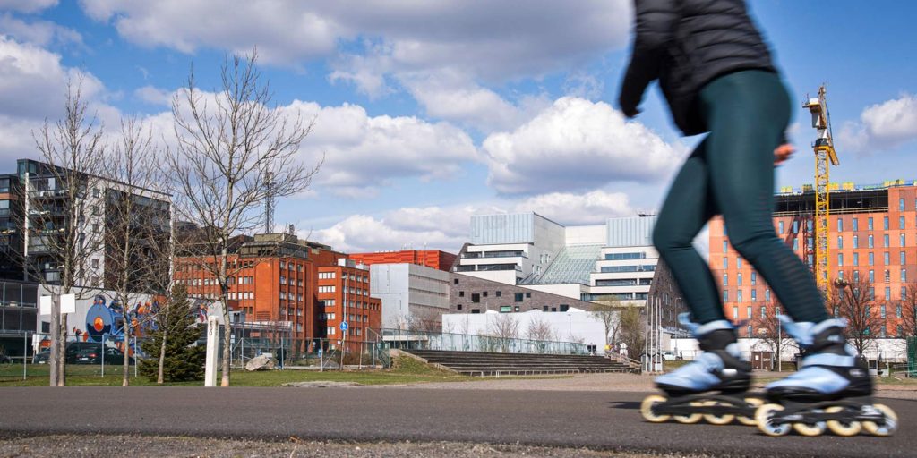 Rullaluistelija liikkuu kaupunkiympäristössä ulkona, taustalla moderneja rakennuksia ja avointa piha-aluetta.
