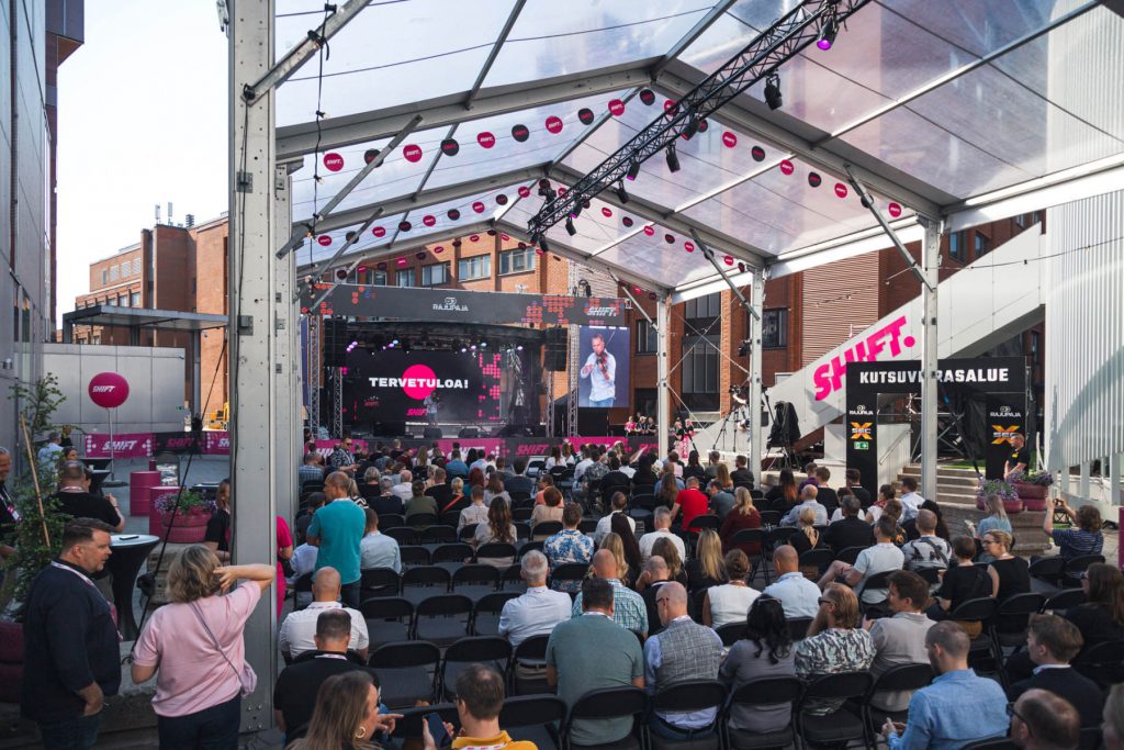 Publiken sitter på stolar och följer en presentation på SHIFT-businessfestivalens huvudscen i ett täckt utomhusutrymme.