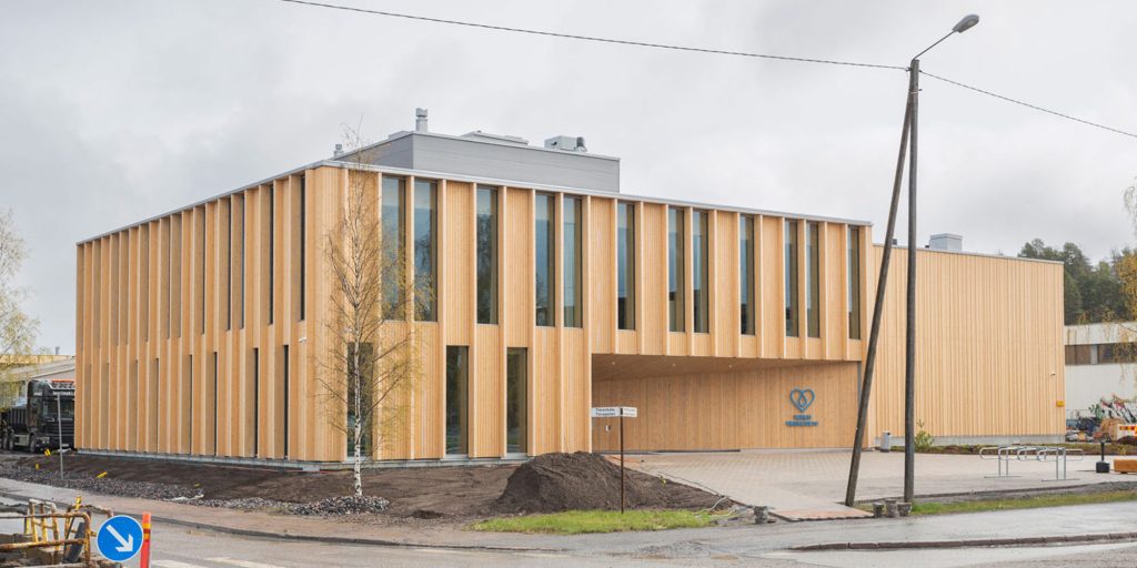 Puuverhoiltu rakennus ulkopuolelta nähtynä, suuret ikkunat ja avoin piha-alue rakennuksen edessä.