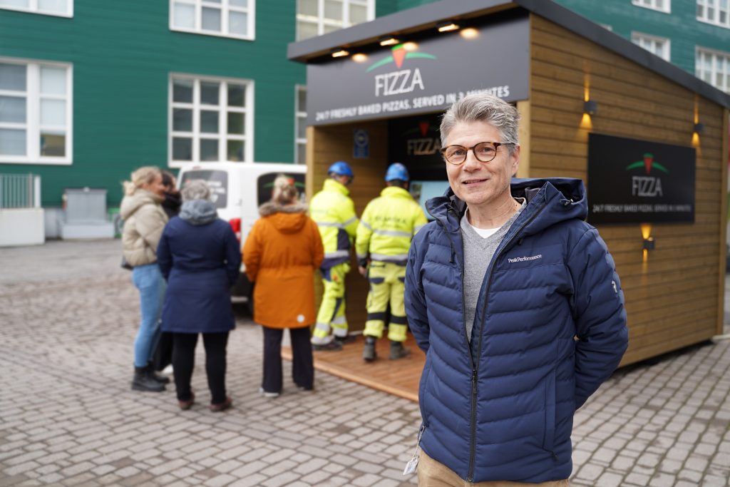Henkilö seisoo ulkona puisen myyntikojun edessä, taustalla muita ihmisiä ja rakennuksia kaupunkiympäristössä.