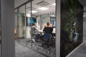 Ett konferensrum med glasväggar i en kontorsmiljö, flera personer sitter vid ett bord och diskuterar, med en skärm och korridor i bakgrunden.