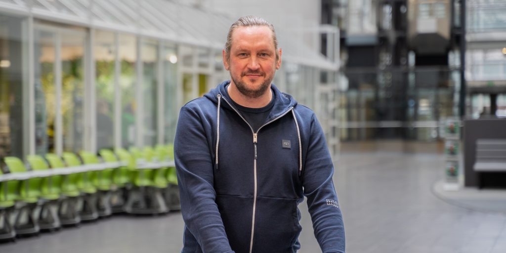 Henkilö seisoo avarassa aulatilassa, taustalla lasiseinäisiä tiloja ja rivissä vihreitä tuoleja.