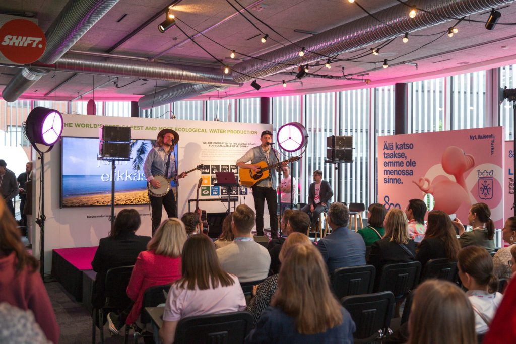 Ett band uppträder på en inomhusscen under SHIFT Business Festival, publiken sitter i rader framför scenen och följer framträdandet.