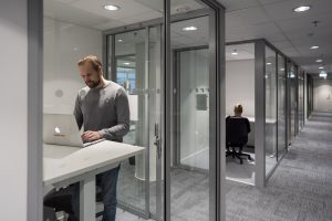 En person arbetar vid en bärbar dator i ett kontorsrum med glasväggar, med en korridor och andra kontorsrum i bakgrunden.